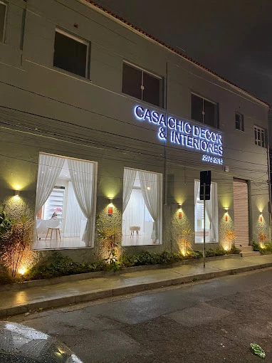 Fachada iluminada da Casa Chic Decor & Interiores à noite, com vitrines elegantes e cortinas sob medida em destaque.
