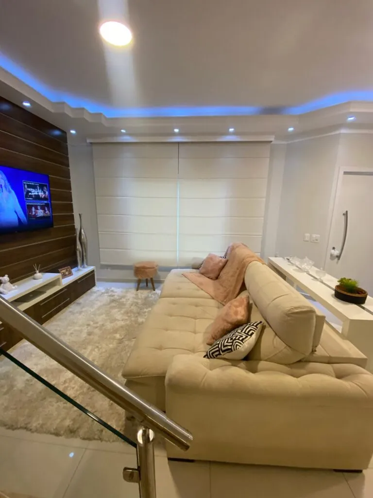 Sala de TV com sofá bege e cortina romana branca, decorada com painel de madeira e iluminação de LED embutida no teto.