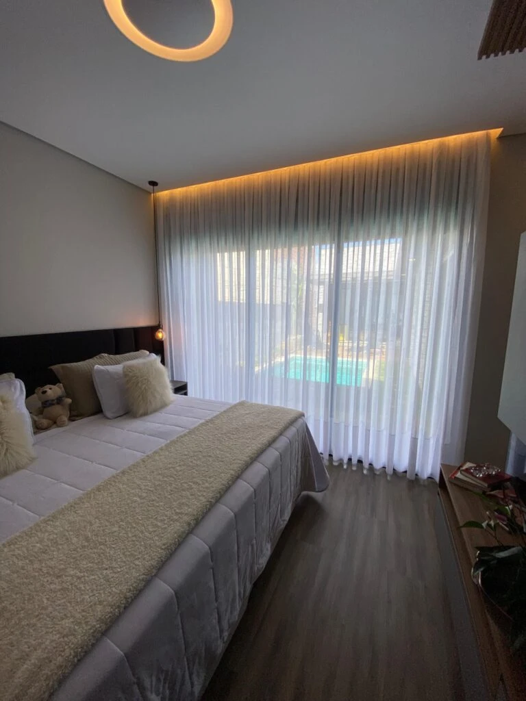 Quarto sofisticado com cama king-size, cortina voil branca iluminada por LED e vista para piscina.